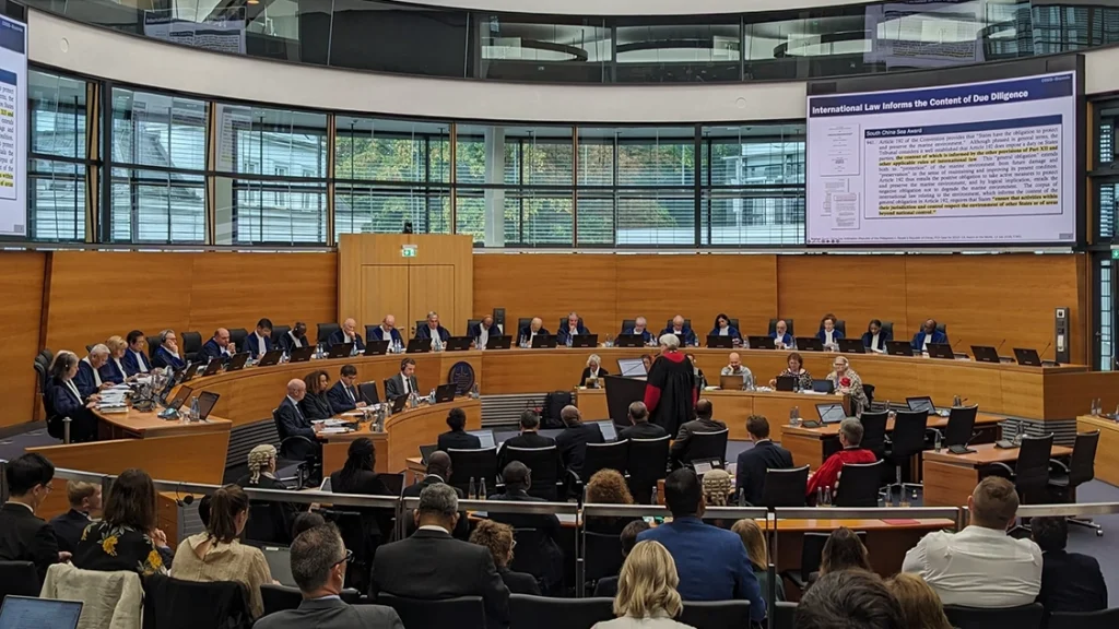 Image of ITLOS plenary, people in suits sitting in a circular room with wooden panelling, and a presentation projected onto a screen (not legible).