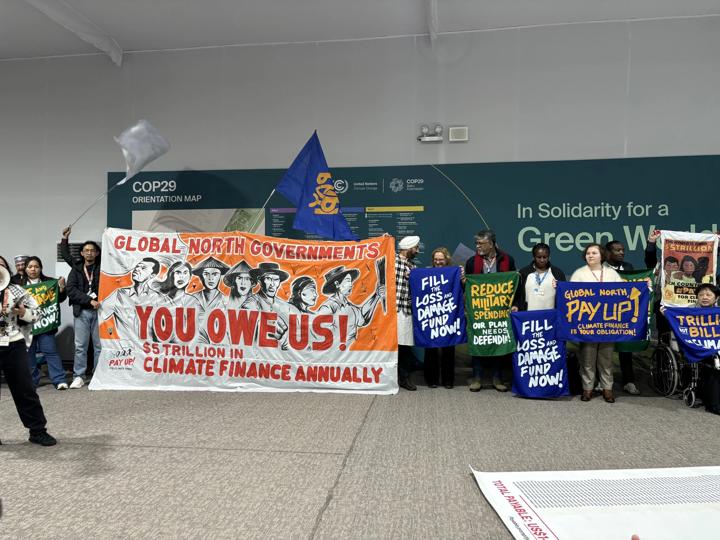 Activists at COP29 in Baku, Azerbaijan, with banners reading:

"Global north governments, you owe us! $5 trillion in climate finance annually."

"Fill the loss and damage fund now!"

"Reduce military spending, our planet needs defending"

"Global north pay up! Climate finance is your obligation!"
