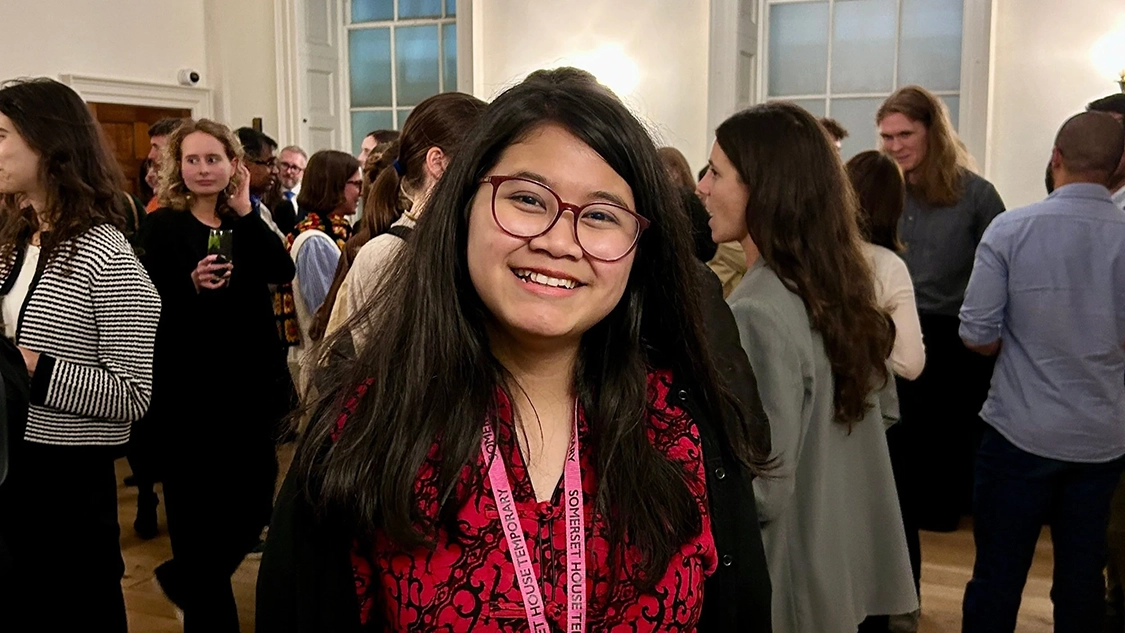 A woman with dark hair and glasses and a red patterned dress smiling. There are a lot of people behind her at a business party. A woman with dark hair and glasses and a red patterned dress smiling. There are a lot of people behind her at a business party.