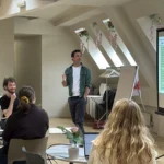 Man in white t-shirt and grey shirt presenting from a projected slide, other staff members sit around tables watching, while smiling and laughing.