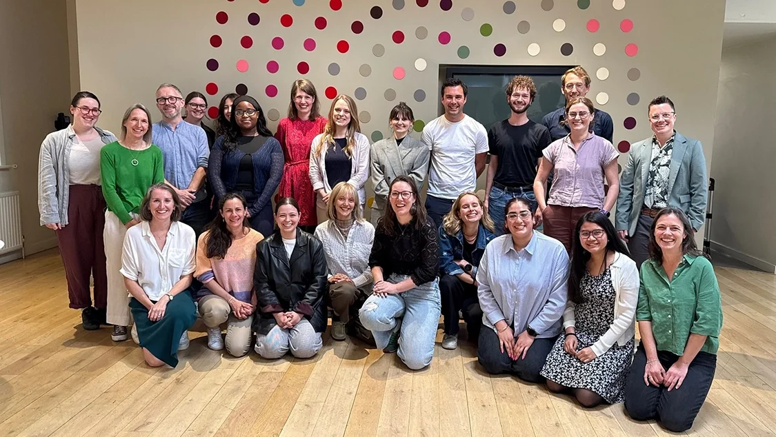 Photo of the Opportunity Green team, 23 staff members in total, some people kneeling, others standing. The floor is wooden and there is a spotted design on the wall behind them.