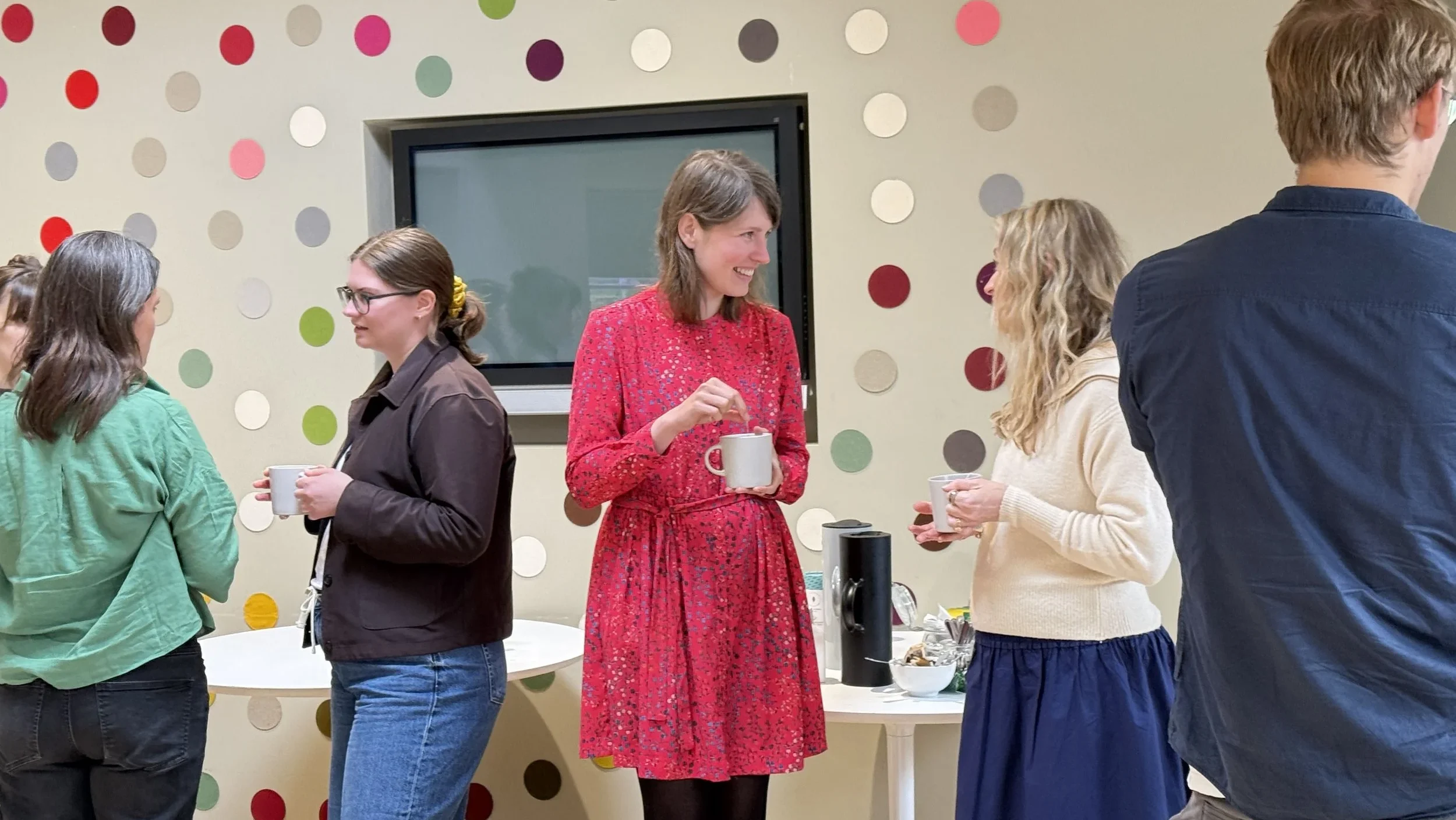 There are three women, the woman on the left wears a brown jumper and her hair tied up and blue jeans. The woman in the middle is holding a mug and wearing a red dress with mid-length hair. She is speaking to the woman on the right who has blond hair, blue trousers and a white jumper. There are coloured circles on the wall in the background.