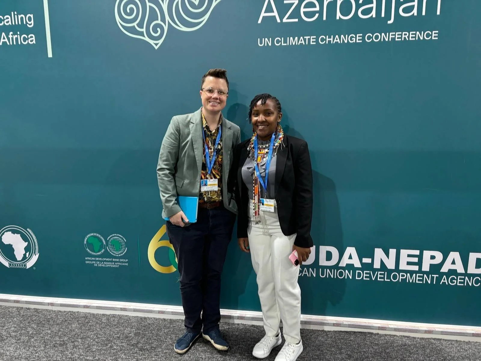 Two people smiling at COP29 in front of a green background. Em Fenton on the left has a grey suit and dark trousers, Elizabeth Wathuti on the right has white trousers and a black jacket.