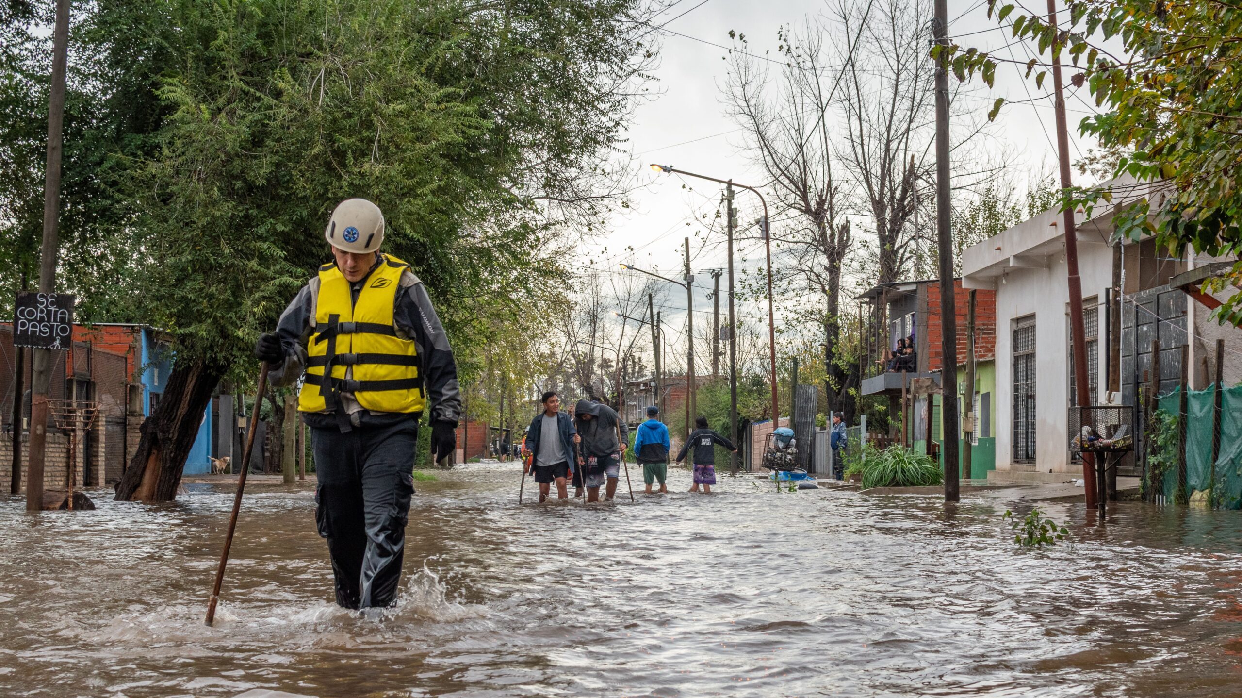 Man in life jacket wading through flooded streets with residents behind him doing the same without safety equipment